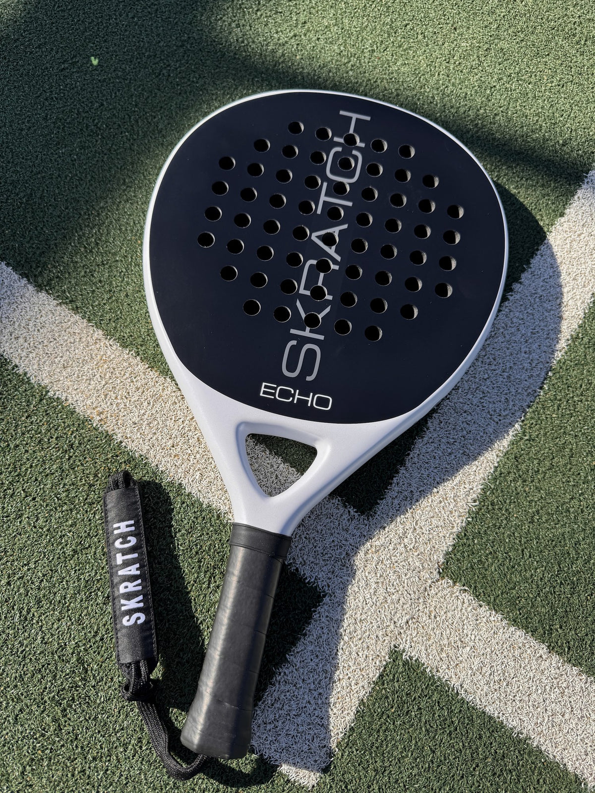 The SKRATCH ECHO padel racket with a matte black carbon face and mist grey pearlescent frame, resting on a green padel court with white boundary lines. The handle is black with a black wrist strap featuring the SKRATCH logo.