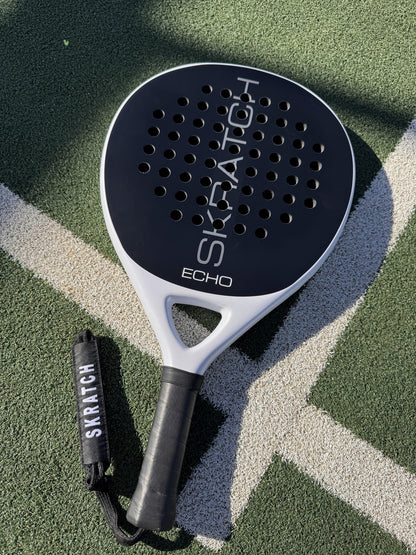 The SKRATCH ECHO padel racket with a matte black carbon face and mist grey pearlescent frame, resting on a green padel court with white boundary lines. The handle is black with a black wrist strap featuring the SKRATCH logo.