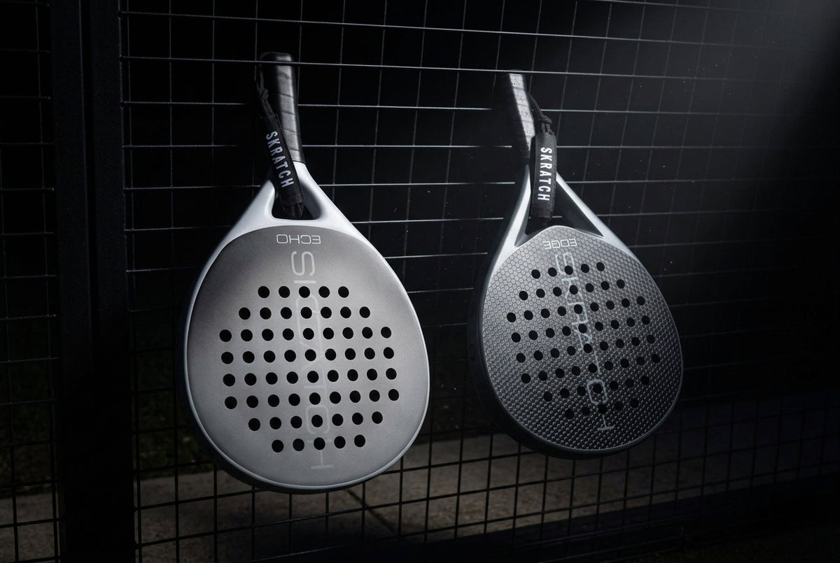 Two SKRATCH padel rackets hanging from a padel court fence, the ECHO model has a matte black face and mist grey pearlescent frame, the EDGE model has a matte black face and charcoal grey pearlescent frame. SKRATCH branding is subtle on both faces.