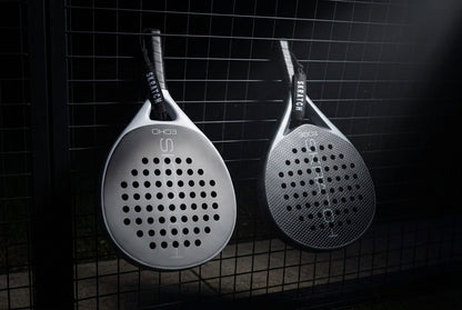 Two SKRATCH padel rackets hanging from a padel court fence, the ECHO model has a matte black face and mist grey pearlescent frame, the EDGE model has a matte black face and charcoal grey pearlescent frame. SKRATCH branding is subtle on both faces.
