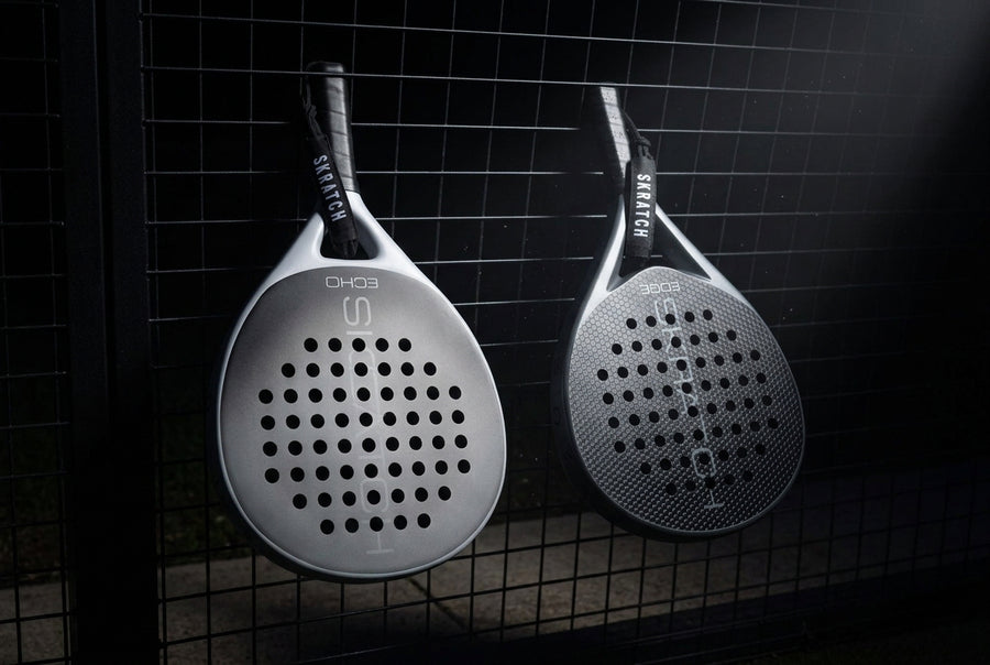Two SKRATCH padel rackets hanging from a padel court fence, the ECHO model has a matte black face and mist grey pearlescent frame, the EDGE model has a matte black face and charcoal grey pearlescent frame. SKRATCH branding is subtle on both faces.