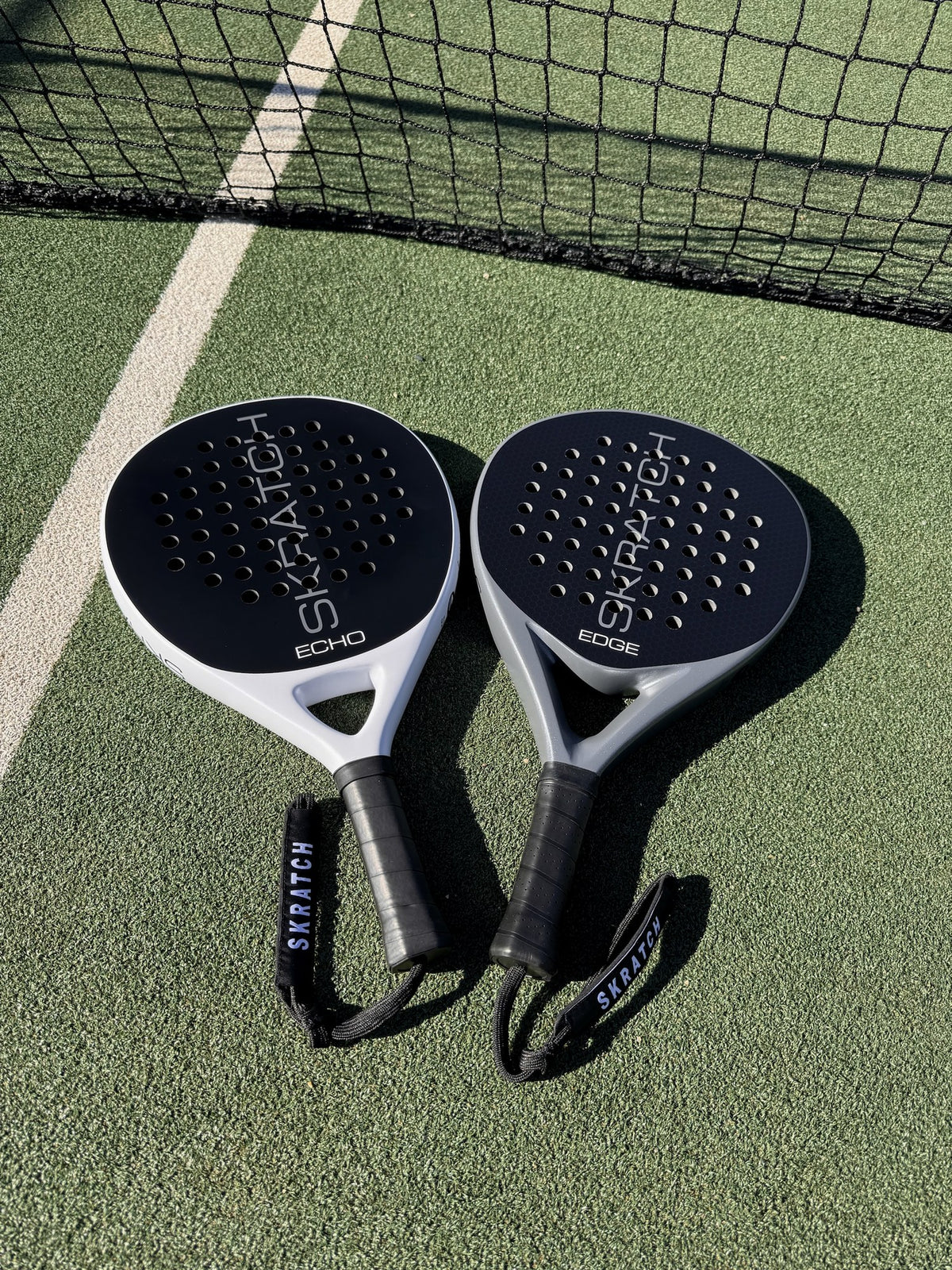 Two SKRATCH padel rackets placed on a green padel court near the net, the ECHO model has a matte black face and mist grey pearlescent frame, the EDGE model has a matte black face and charcoal grey pearlescent frame. SKRATCH branding is subtle on both faces.