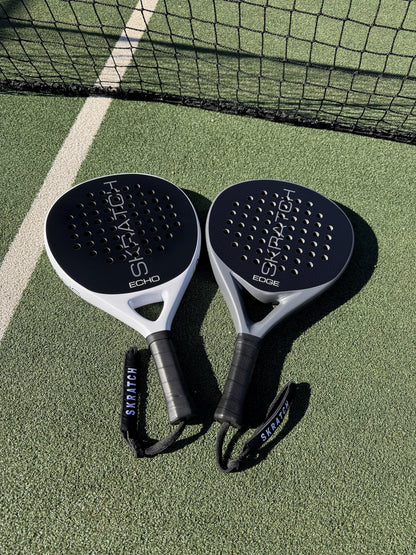 Two SKRATCH padel rackets placed on a green padel court near the net, the ECHO model has a matte black face and mist grey pearlescent frame, the EDGE model has a matte black face and charcoal grey pearlescent frame. SKRATCH branding is subtle on both faces.